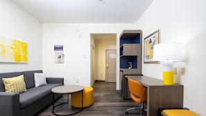 A modern living room with a gray sofa, yellow ottoman, and a round coffee table. There is a desk with a chair and a lamp. The room has a wooden floor and white walls with artwork. - Comfort Suites Oil Center (Lafayette)