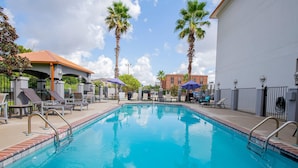 A swimming pool with lounge chairs and umbrellas, surrounded by palm trees and a building with a red roof. - Comfort Suites Oil Center (Lafayette)