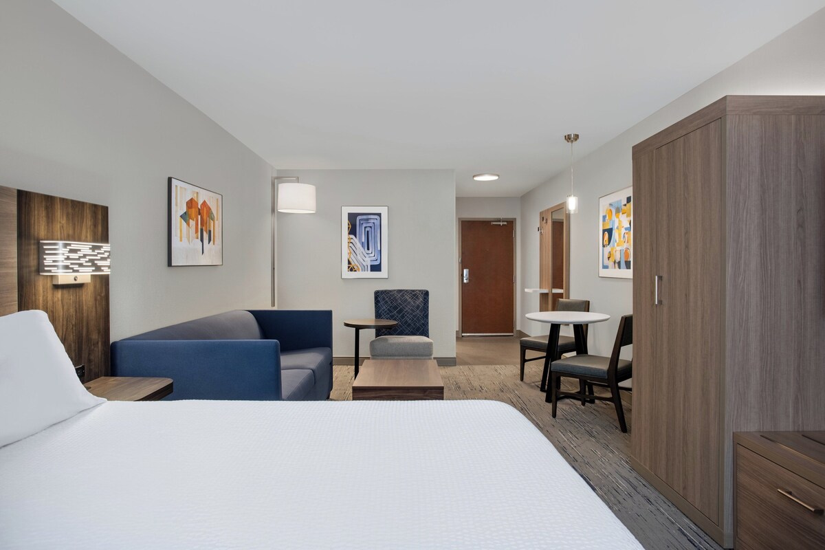 A modern hotel room with a large bed, a blue sofa, a small table, a chair, and a wardrobe.