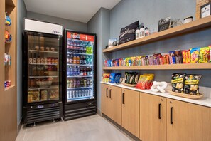 A vending area with two refrigerators stocked with snacks and drinks, and shelves above containing more snacks and a few bottles. - Holiday Inn Express & Suites Terre Haute by IHG (Terre Haute)