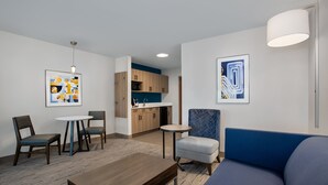 Una sala moderna con un sofá azul, una mesa de centro de madera y una pequeña mesa redonda con sillas. En el fondo, se aprecia una kitchenette con armarios de madera y una encimera azul.