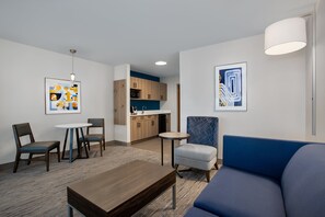 A modern living room with a blue sofa, a wooden coffee table, and a small round table with chairs. There is a kitchenette in the background with wooden cabinets and a blue countertop. - Holiday Inn Express & Suites Terre Haute by IHG (Terre Haute)