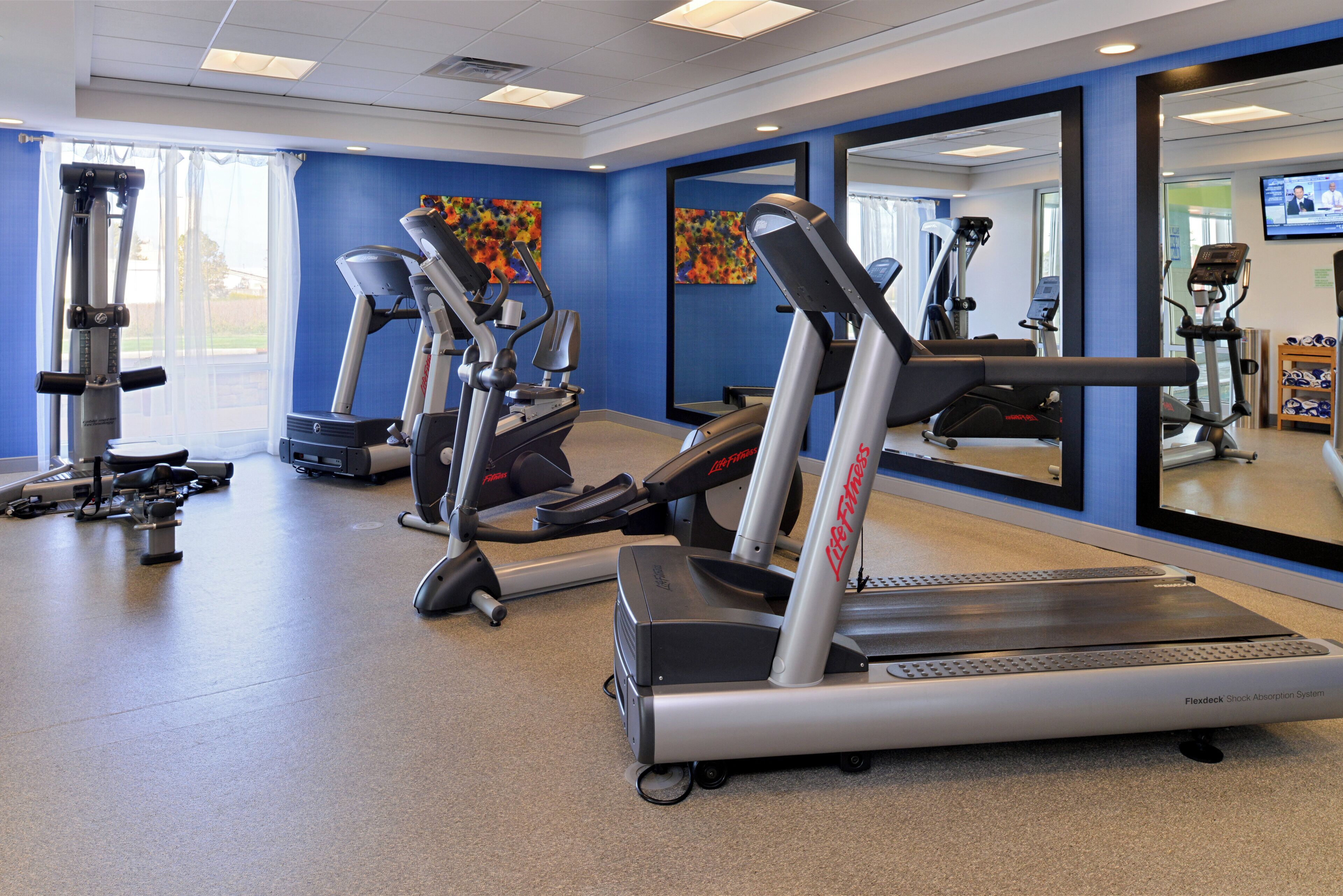 A gym with various cardio machines, a mirrored wall, and a large window.