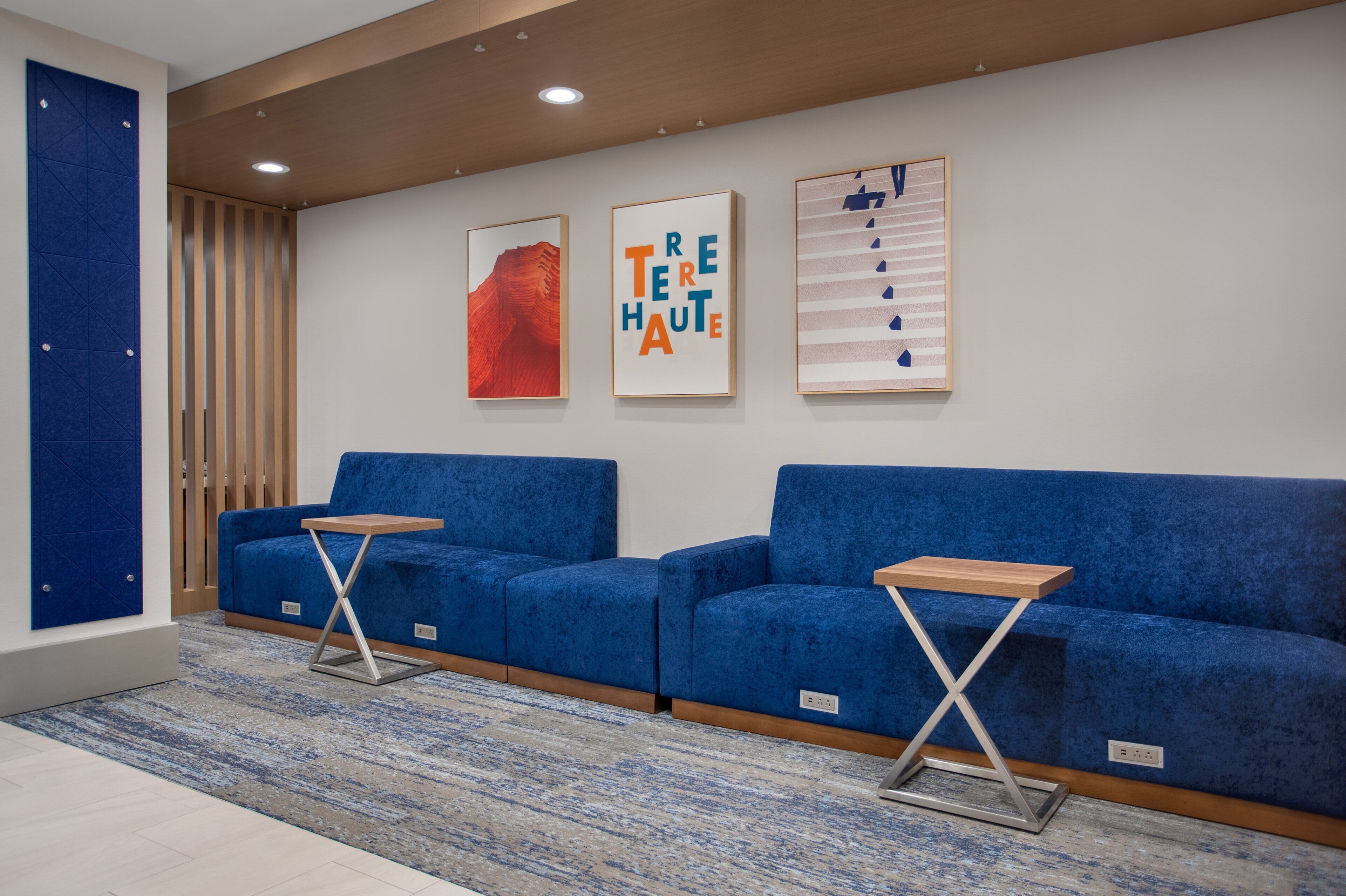 A modern waiting area with two blue sofas, a wooden table, and three framed artworks on the wall.