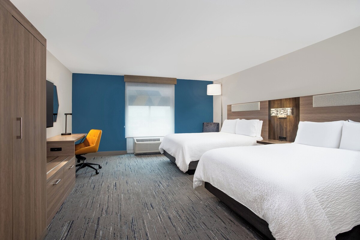 A hotel room with two beds, a desk, a chair, and a large window.
