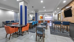 A modern hotel dining area with multiple tables and chairs, a large TV screen, and a central pillar with a geometric design. - Holiday Inn Express & Suites Amarillo West by IHG (Amarillo)