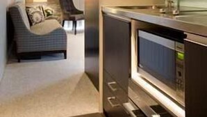 Una cocina moderna con electrodomésticos de acero inoxidable, una zona de estar con sofá y sillones, y vista a un salón con una lámpara y cortinas.