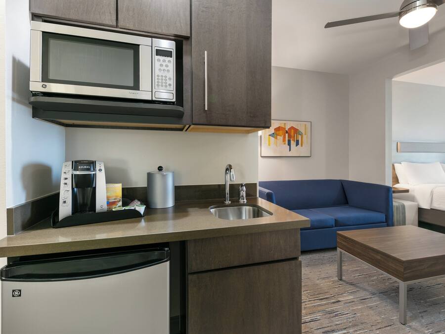 A compact kitchen with a microwave, coffee maker, and sink. A blue sofa and a bed with a headboard are visible in the adjacent living area.