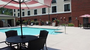 A pool area with a table and chairs, a poolside umbrella, and a brick building in the background.