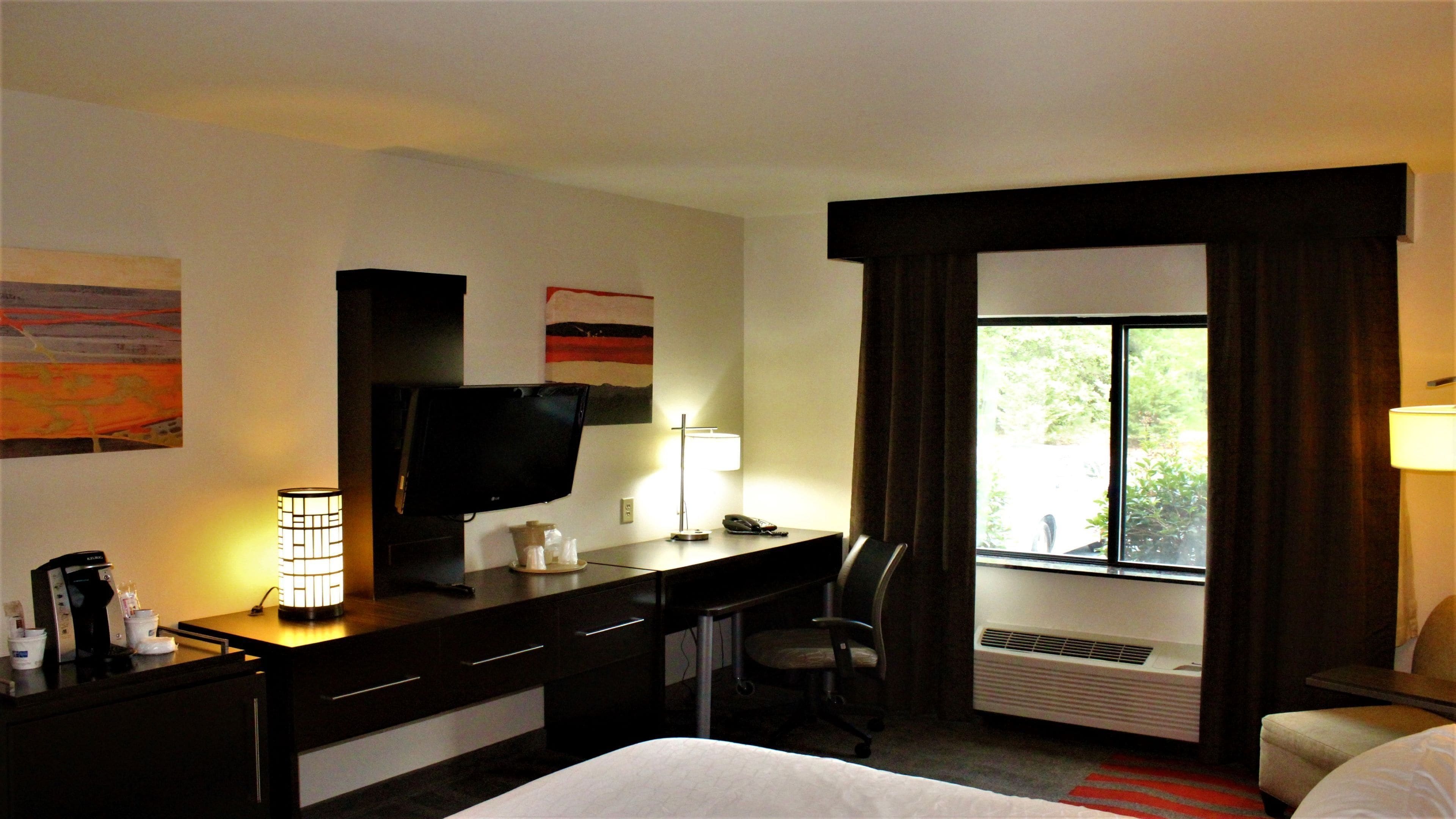 A hotel room with a bed, a desk, a chair, a television, a lamp, and a window with curtains.