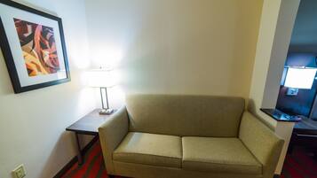 Um sofá bege num quarto de hotel com um tapete listrado em vermelho e preto, um quadro abstrato emoldurado na parede e uma pequena mesa com uma luminária.
