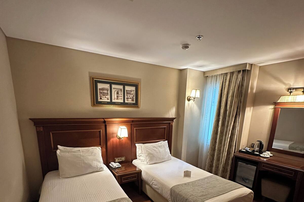 A hotel room with two single beds, a desk, a chair, a nightstand with a lamp, and a picture frame on the wall.