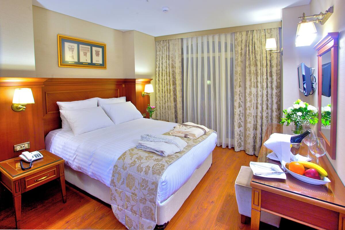 A hotel room with a bed, bedside lamps, a bedside table with a phone, a desk with a mirror, and a fruit bowl.