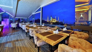 A restaurant with plush seating, large windows, and a view of a mosque at night. - Perula Hotel & SPA (Istanbul)