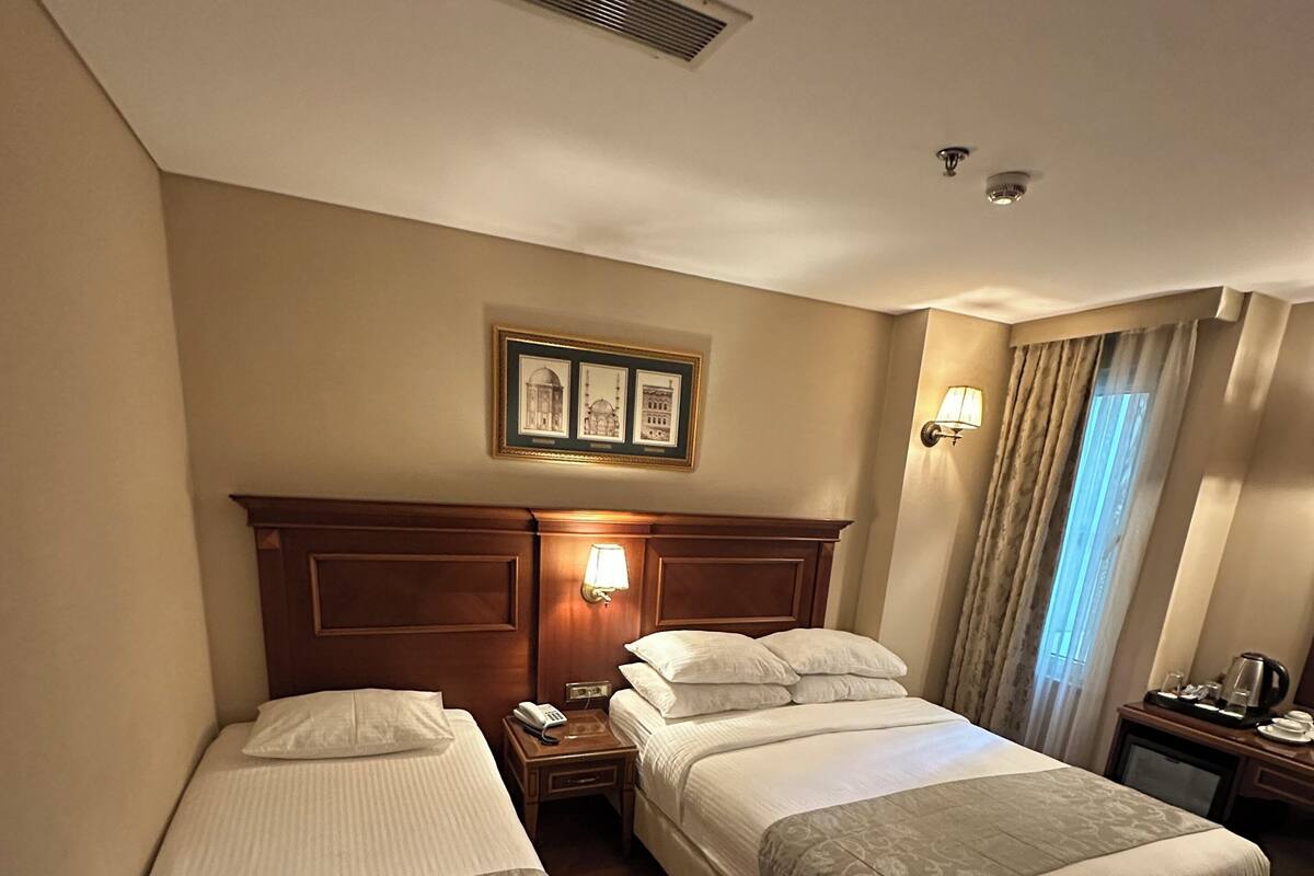 A hotel room with two single beds, a wooden headboard, a nightstand with a lamp, a framed picture on the wall, and a window with curtains.