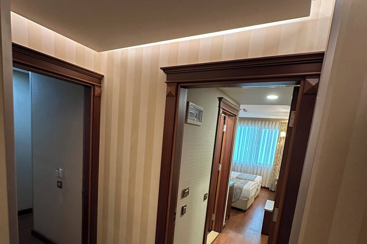 A hotel hallway with patterned carpet, wooden doors, and a visible bedroom with a bed and curtains.