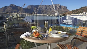 A table with a meal, a glass of wine, and a bowl of salad, overlooking a cityscape with mountains and construction cranes.