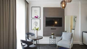 A modern living room with a flat-screen TV, a grey armchair, a round table, and a small side table with a vase of flowers.