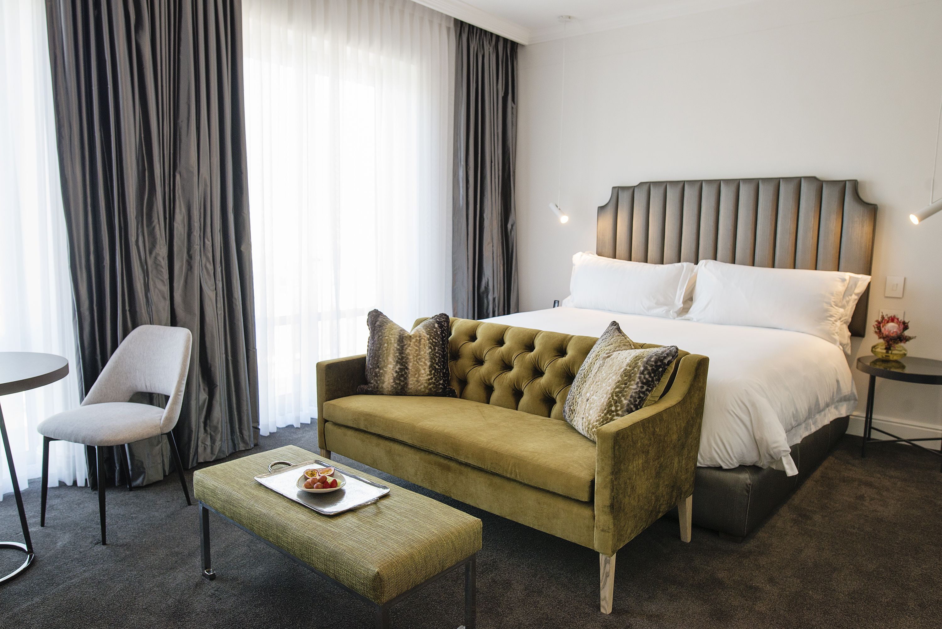 A hotel room with a bed, a green sofa, a small table with a tray of fruit, a chair, and a nightstand with a lamp.