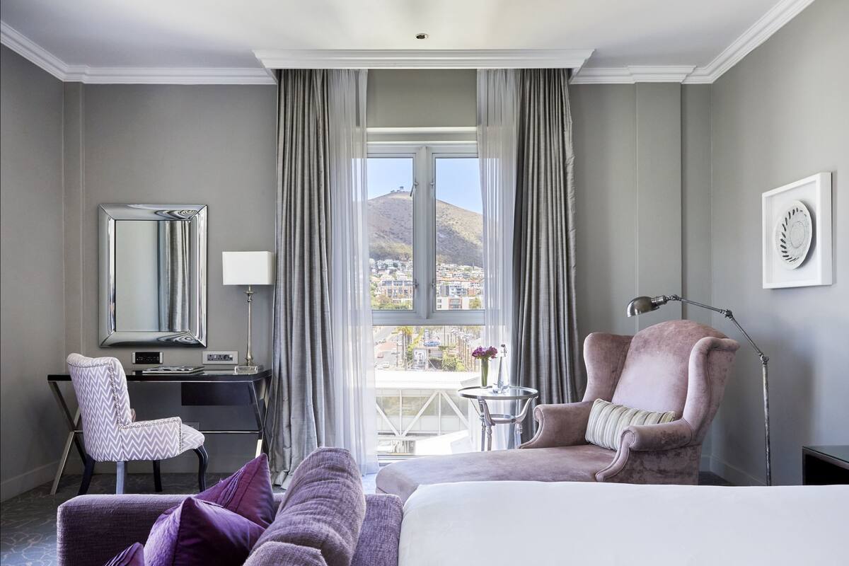 A hotel room with a bed, a desk, a chair, a pink armchair, and a view of a cityscape through large windows.