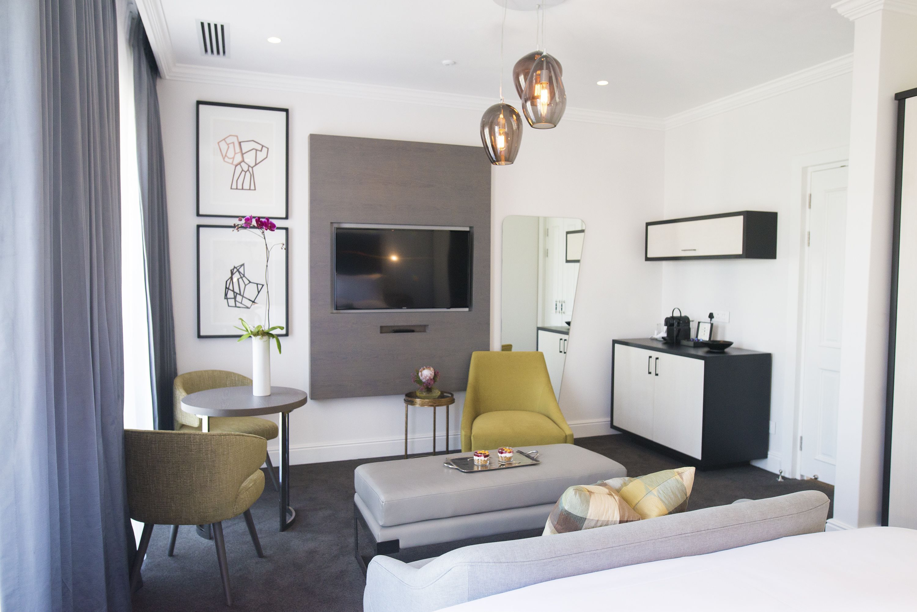 Ein modernes Hotelzimmer mit grauem Sofa, einem kleinen runden Tisch, einem gelben Sessel, einem grauen Hocker, einem Flachbildfernseher, einer schwarz-weißen Minibar und einem großen Fenster mit grauen Vorhängen.