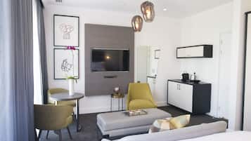 A modern hotel room with a grey sofa, a small round table, a yellow armchair, a grey ottoman, a flat-screen TV, a black and white minibar, and a large window with grey curtains.