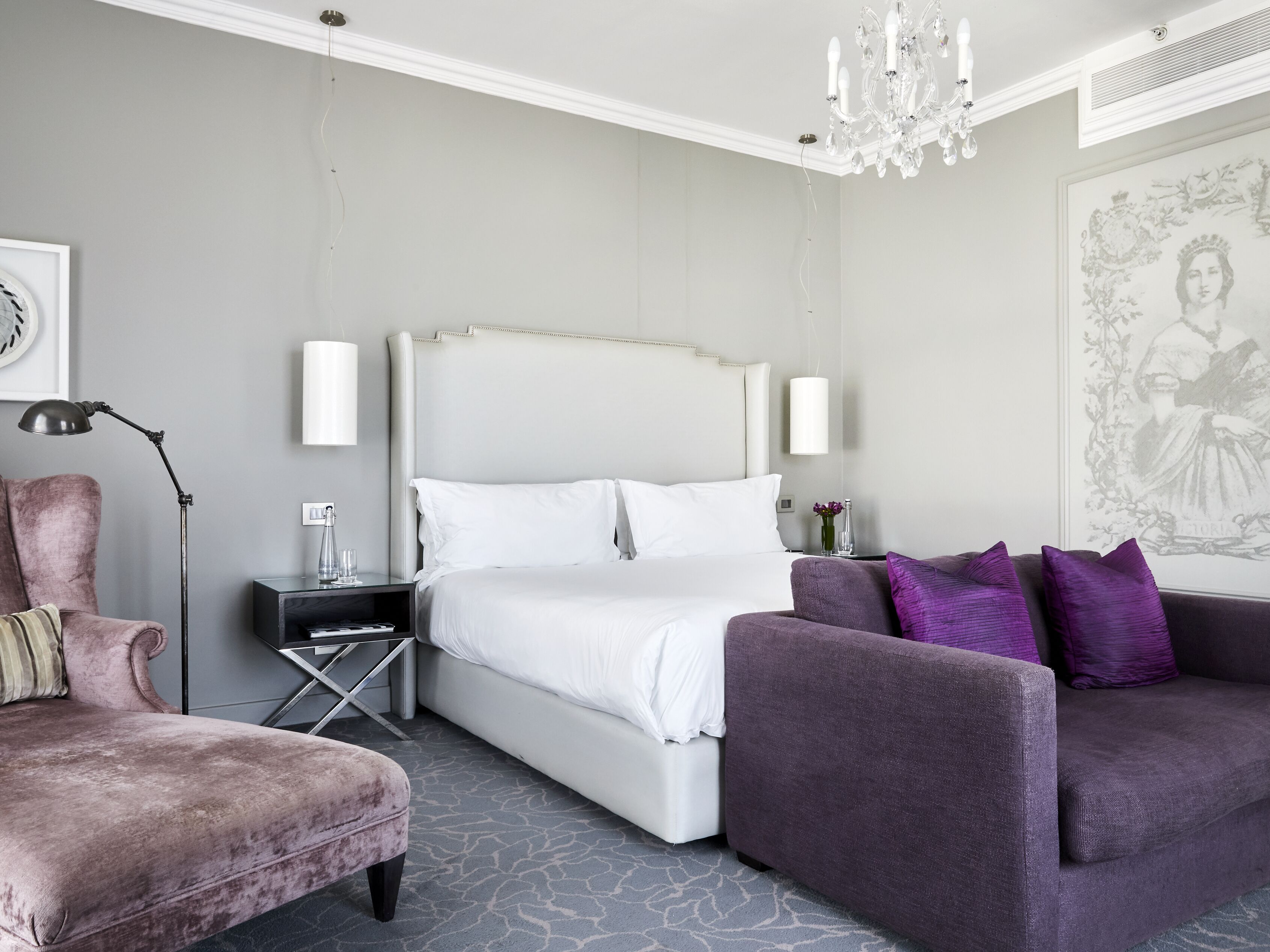 A modern hotel room with a large bed, a seating area with a purple sofa, a pink armchair, and a chandelier.