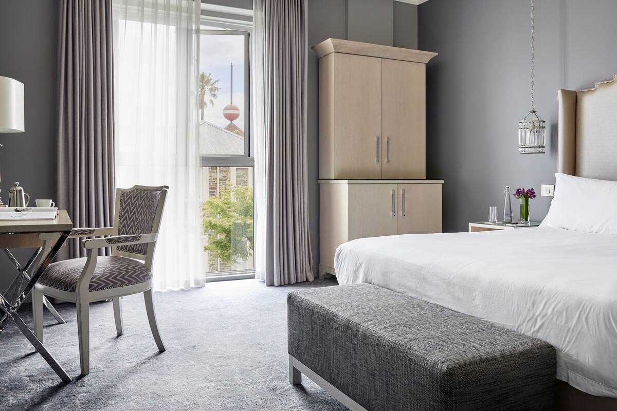 A hotel room with a bed, a desk with a chair, a wardrobe, and a view of the outside.