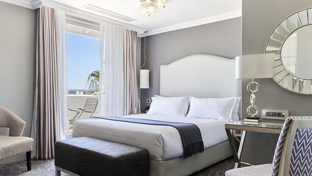A hotel room with a large bed, a desk, a chair, a balcony view, and a chandelier.