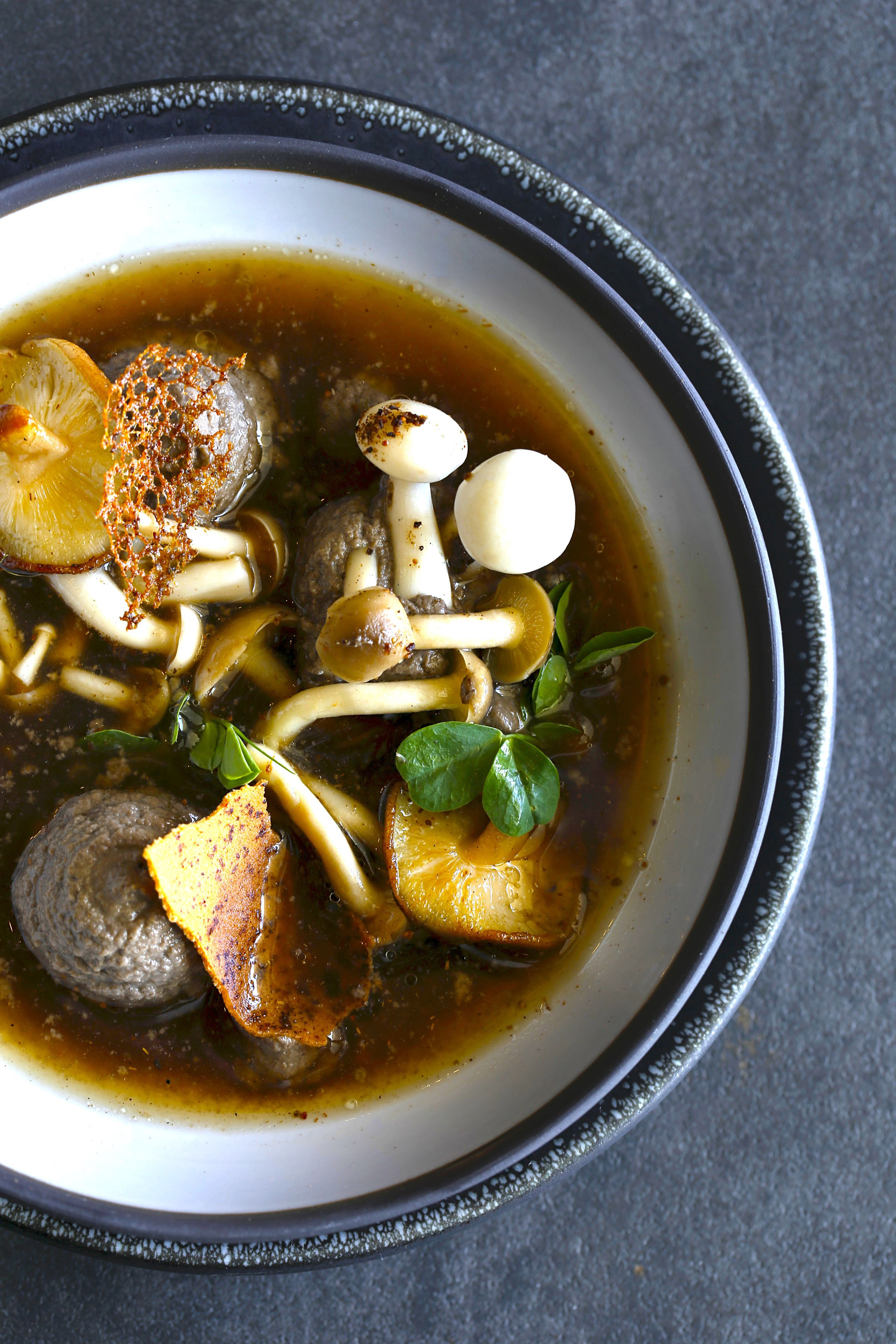 A bowl of soup with various ingredients like mushrooms, herbs, and possibly meatballs, served in a dark bowl with a decorative rim.