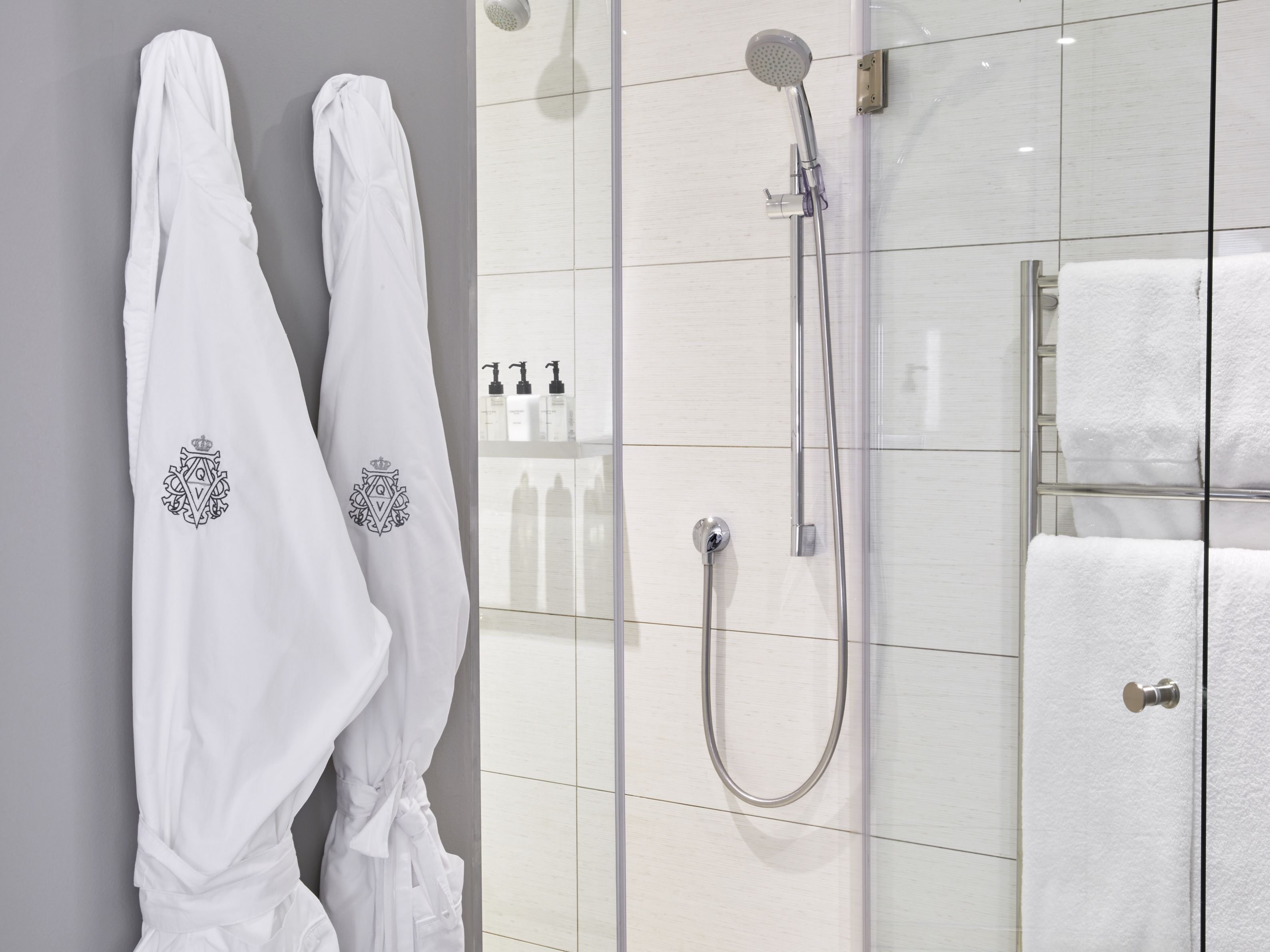 A bathroom with two white robes hanging on a wall, a glass shower enclosure, and a towel rack with white towels.