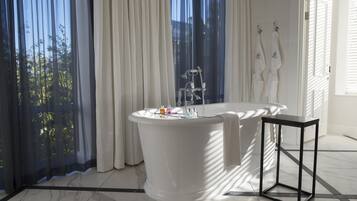 A modern bathroom with a freestanding bathtub, a small table, and a view of greenery through sheer curtains.