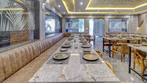 A spacious dining area with a long table set for a meal, cushioned seating along the walls, and a modern ceiling design. - Hotel Anand Villa (Agra)