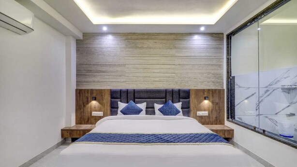 A modern bedroom with a large bed, wooden headboard, two bedside tables, and a glass wall with a view. - Hotel Anand Villa (Agra)