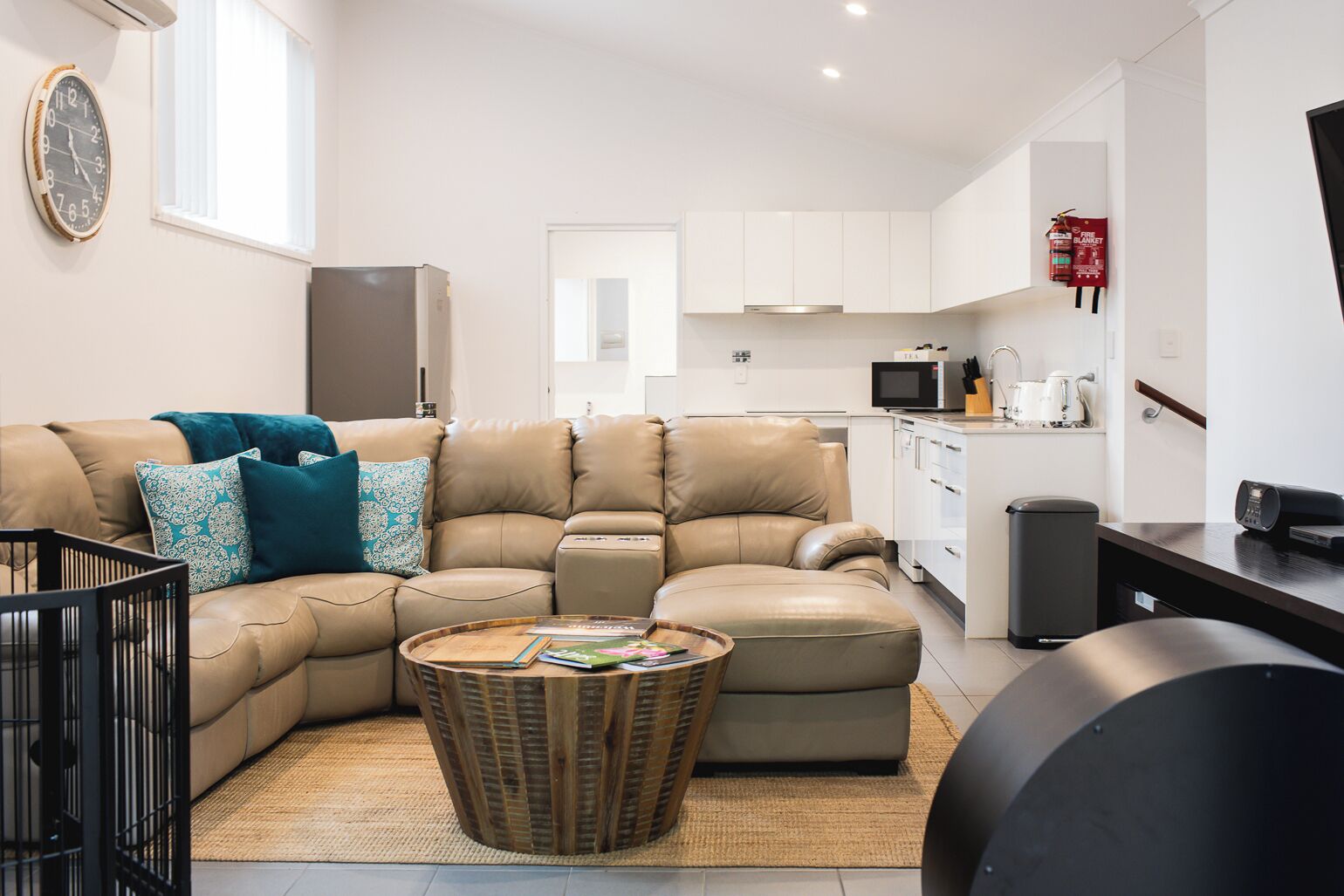 Un salon moderne avec un canapé beige, une table basse et un coin cuisine doté de meubles blancs et d’appareils électroménagers.