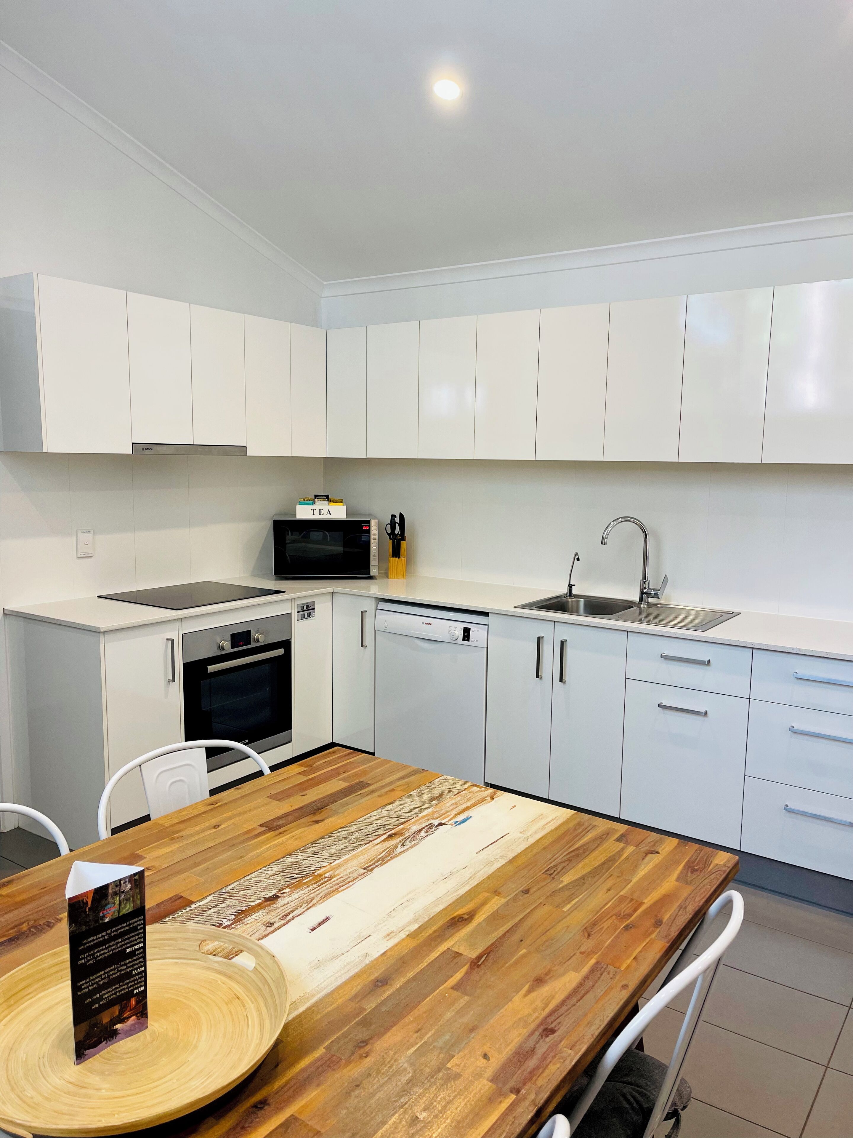 A modern kitchen with white cabinets, a microwave, oven, and dishwasher. There is a wooden dining table with chairs and a placemat.