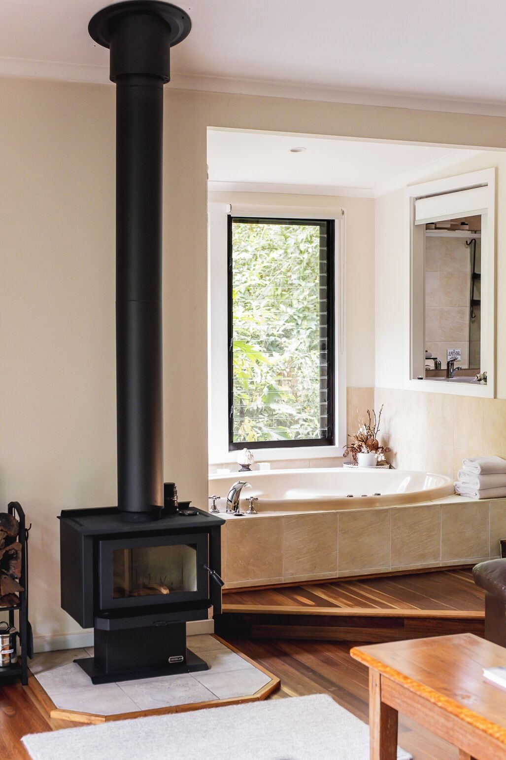 Una sala de estar moderna con un estufa de leña negra, un ventanal con vistas a zonas verdes, un baño con bañera y espejo, y piso de madera.