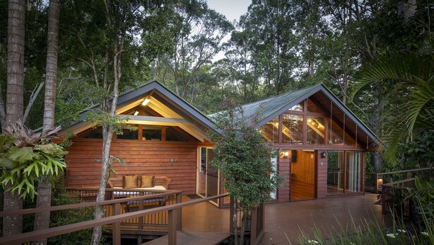 Dos cabañas de madera con paredes de vidrio, conectadas por un puente de madera, rodeadas de un bosque denso.