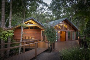 Deux cabanes en bois aux murs de verre, reliées par un pont de bois, et entourées d’une forêt dense.