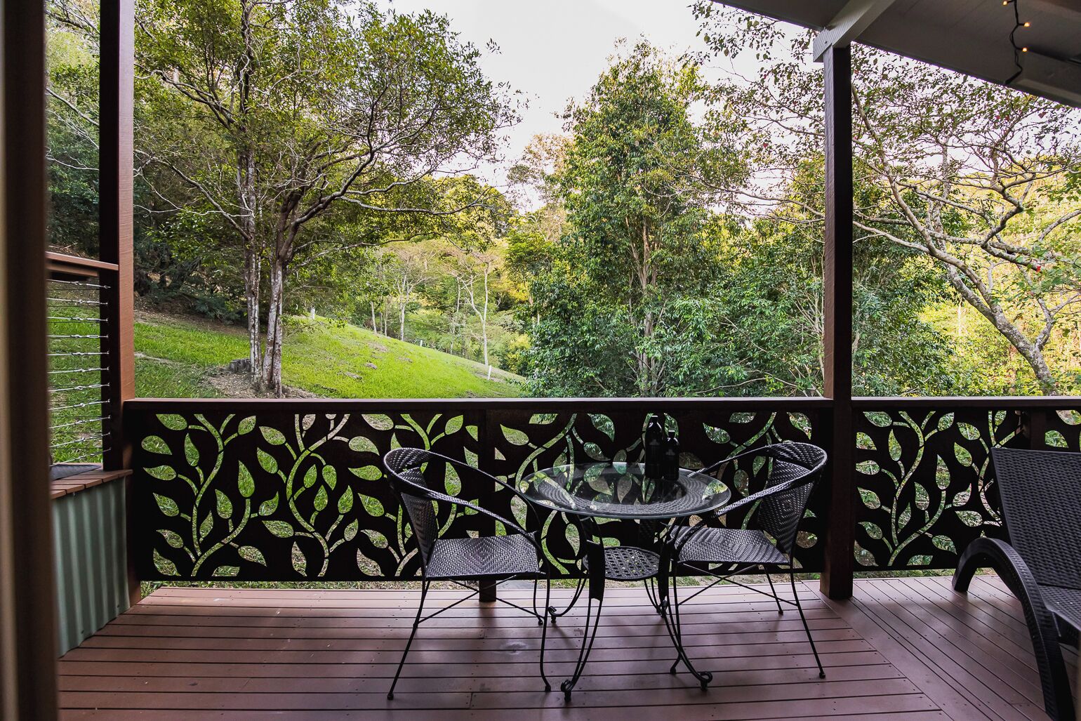 Un espacio al aire libre cubierto con una mesa y sillas, rodeado por una terraza de madera y una barandilla decorativa con motivos de hojas.