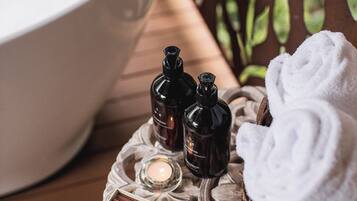A bathroom vanity with a white sink, two dark bottles, a lit candle, a rolled white towel, and a card with text.