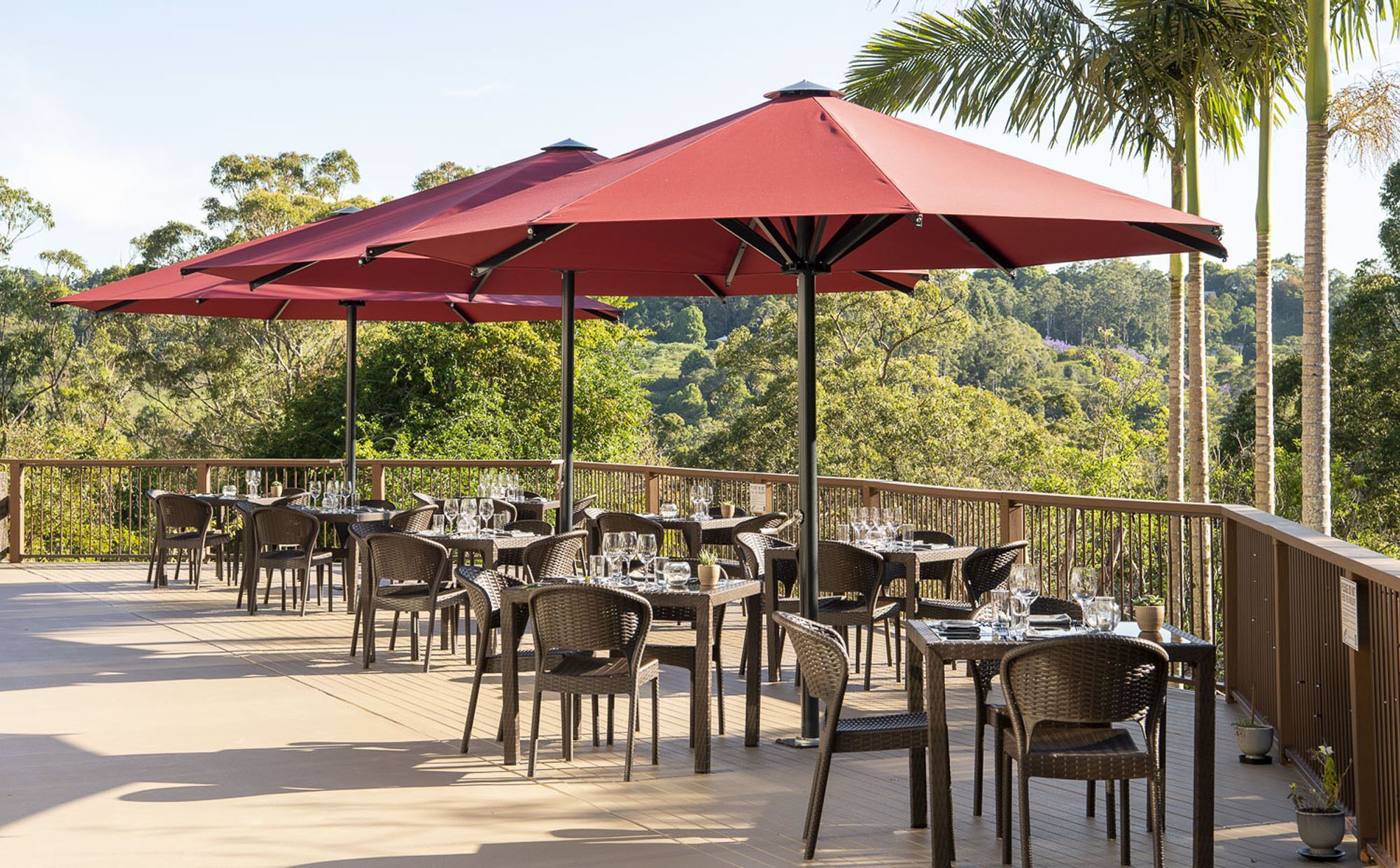 An outdoor dining area with tables and chairs, red umbrellas, and a view of a forested landscape.