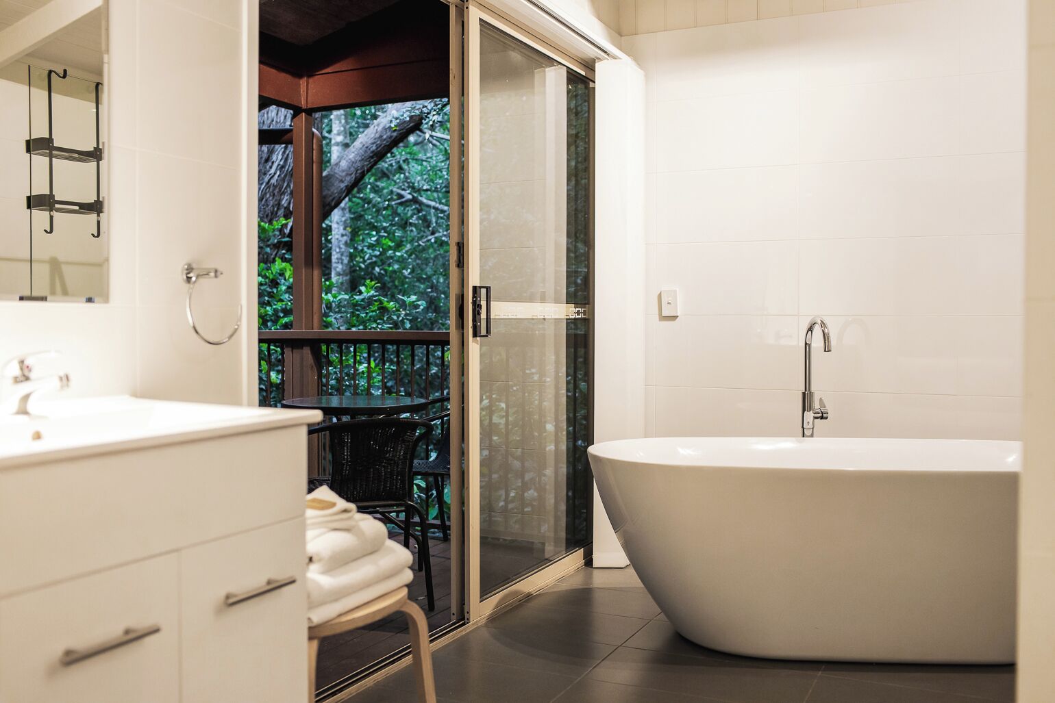 A modern bathroom with a freestanding bathtub, a vanity with a mirror, and a glass door leading to an outdoor area with a view of trees.