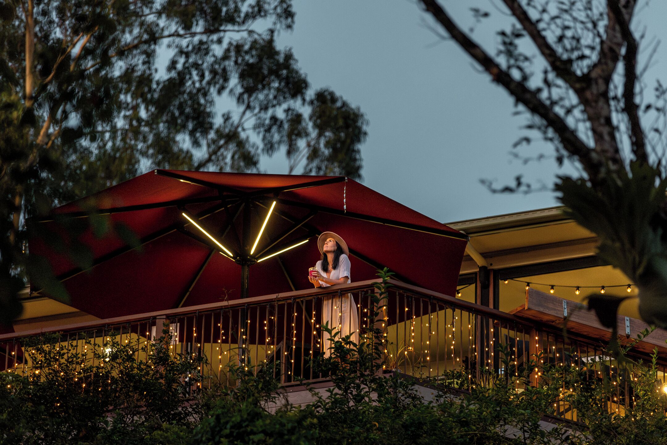 A person standing on a balcony with a red umbrella, surrounded by greenery and string lights.