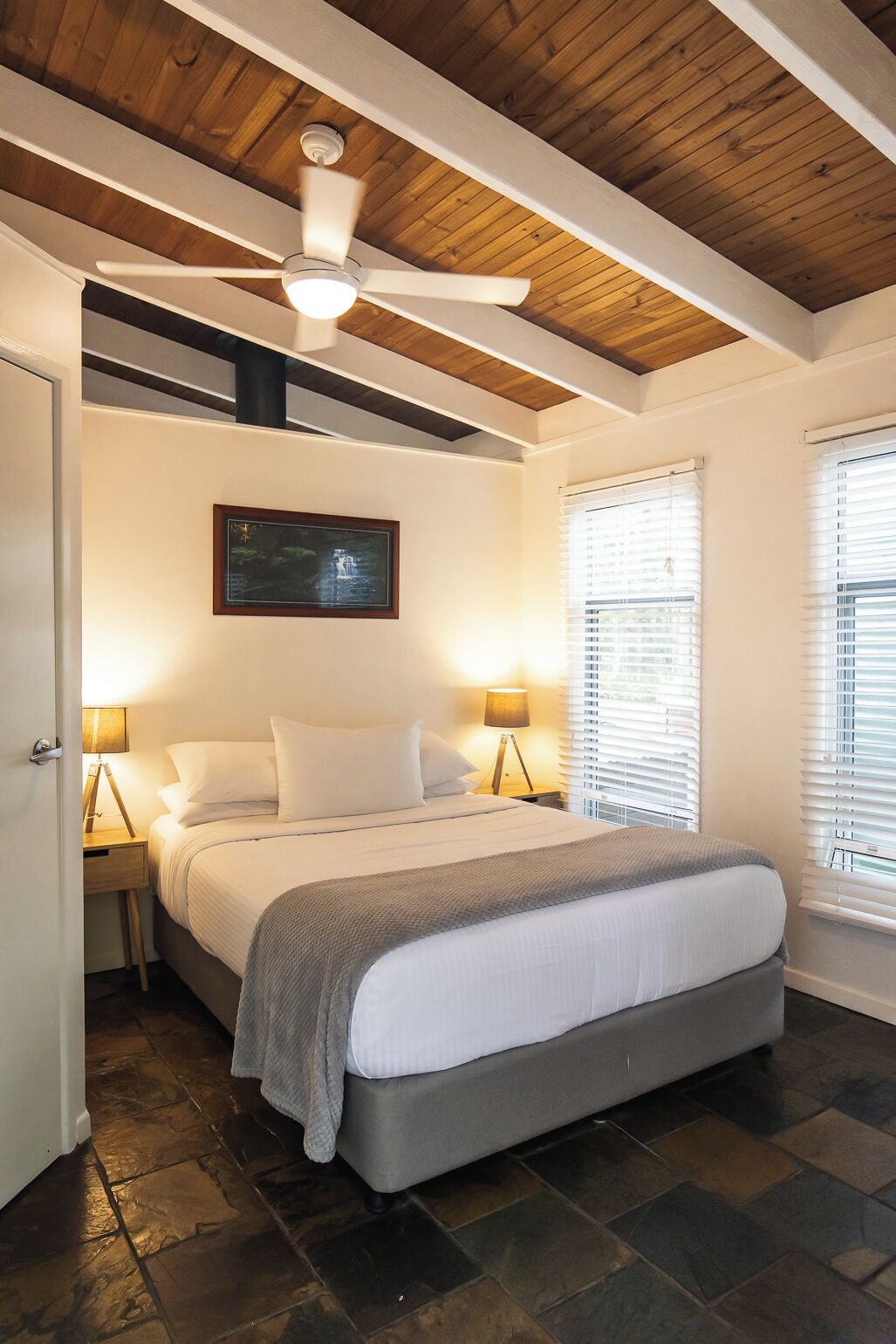 A bedroom with a bed, two bedside lamps, a ceiling fan, a framed picture on the wall, and a window with blinds.