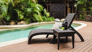 Zona de piscina con una silla de salón y una mesa, rodeada de abundante vegetación y una terraza de madera.