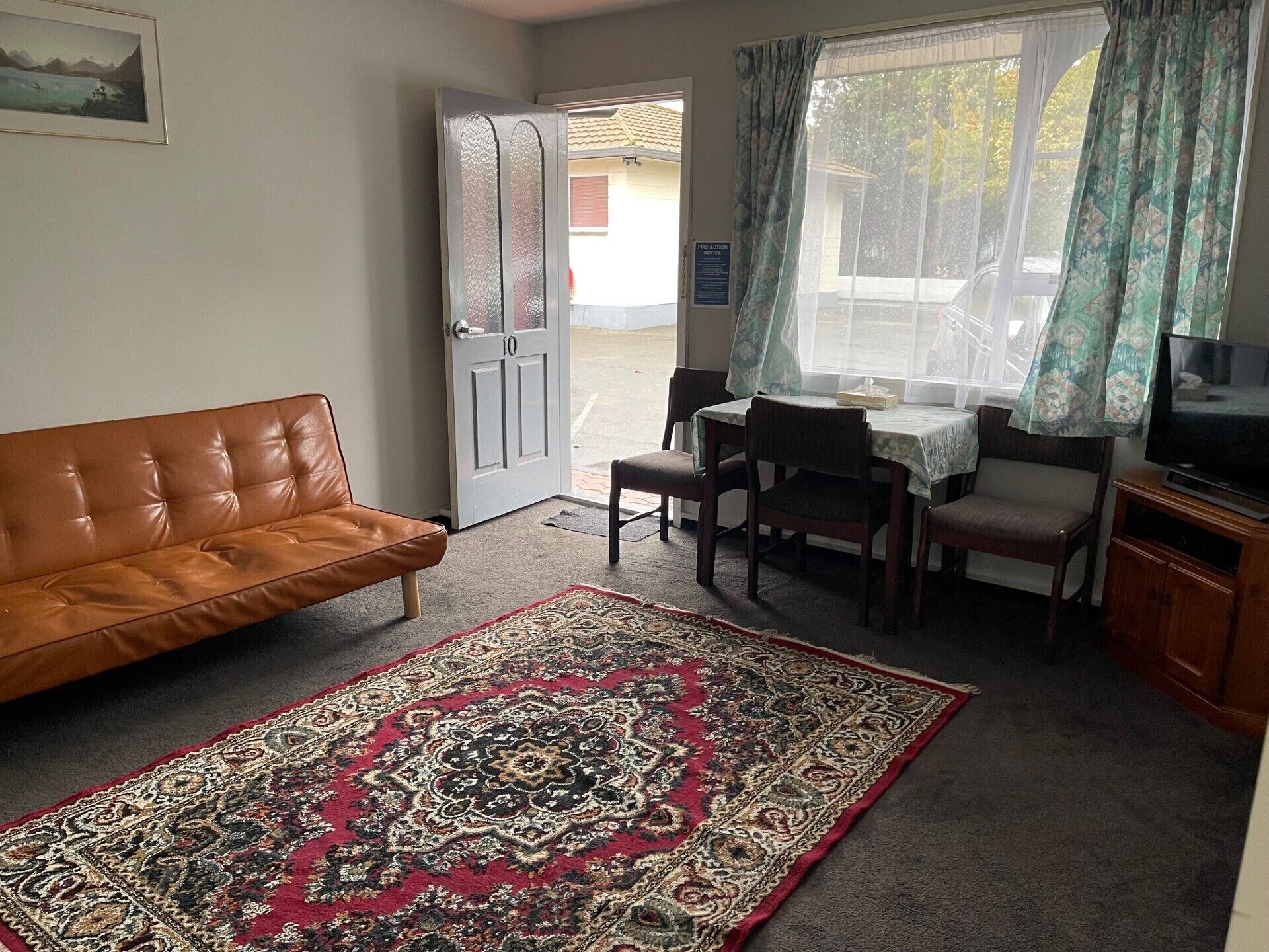Un salon avec un canapé en cuir marron, un tapis à motifs, une table à manger avec des chaises, un téléviseur et une porte donnant sur l’extérieur.