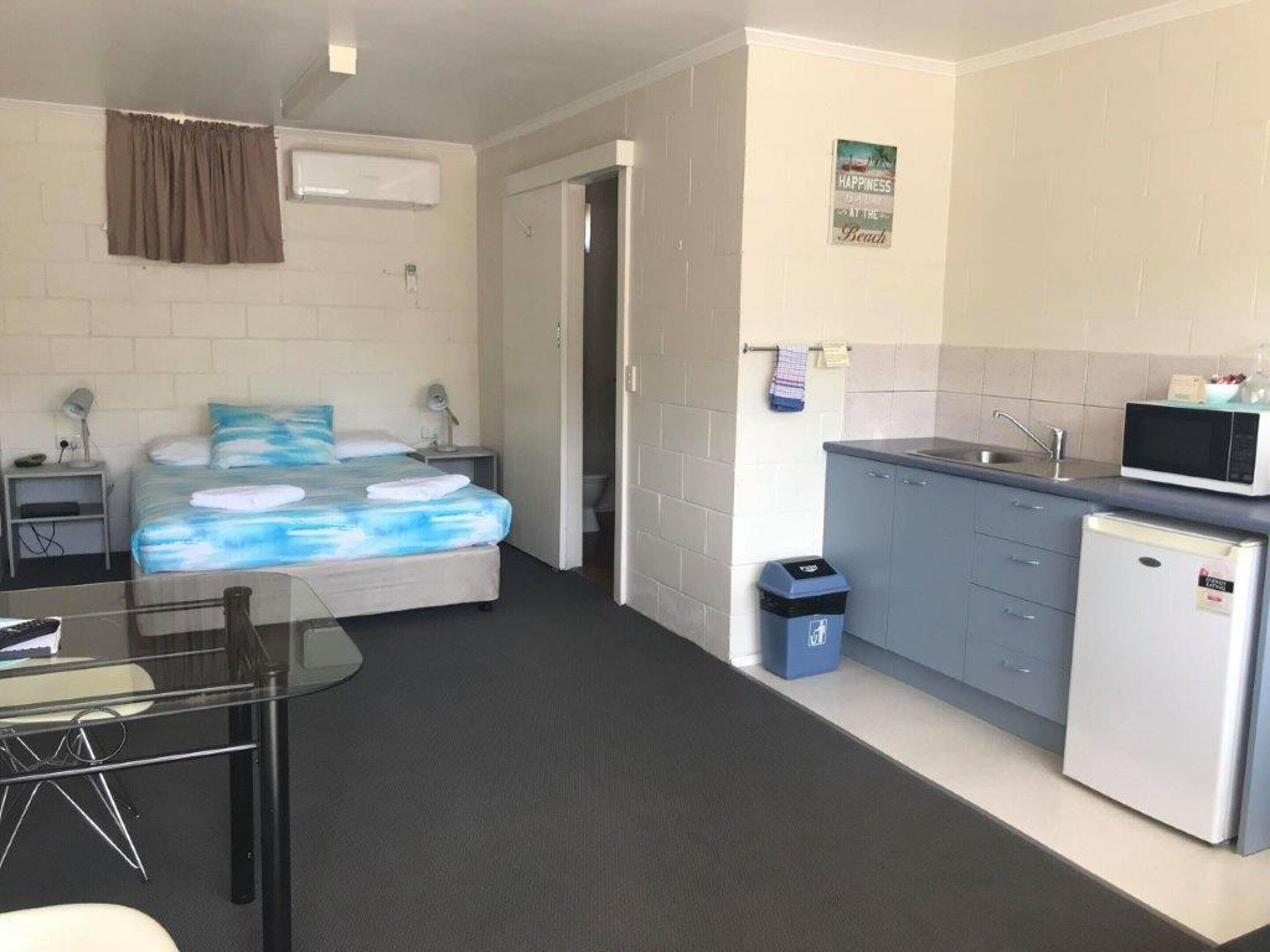 A compact hotel room with a bed, bedside table, lamp, and a small kitchenette with a microwave and refrigerator.