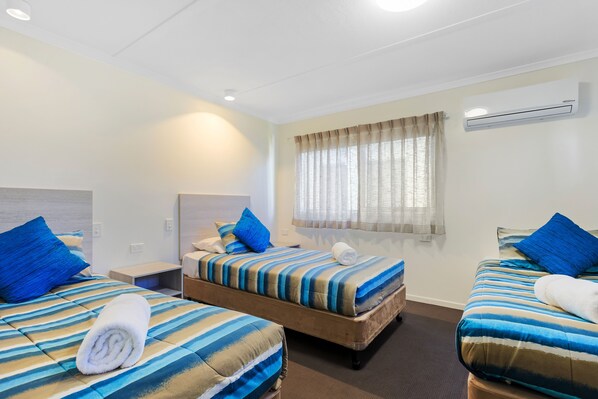 A hotel room with two beds, each with striped bedding and blue pillows, a window with sheer curtains, and a ceiling-mounted air conditioner.
