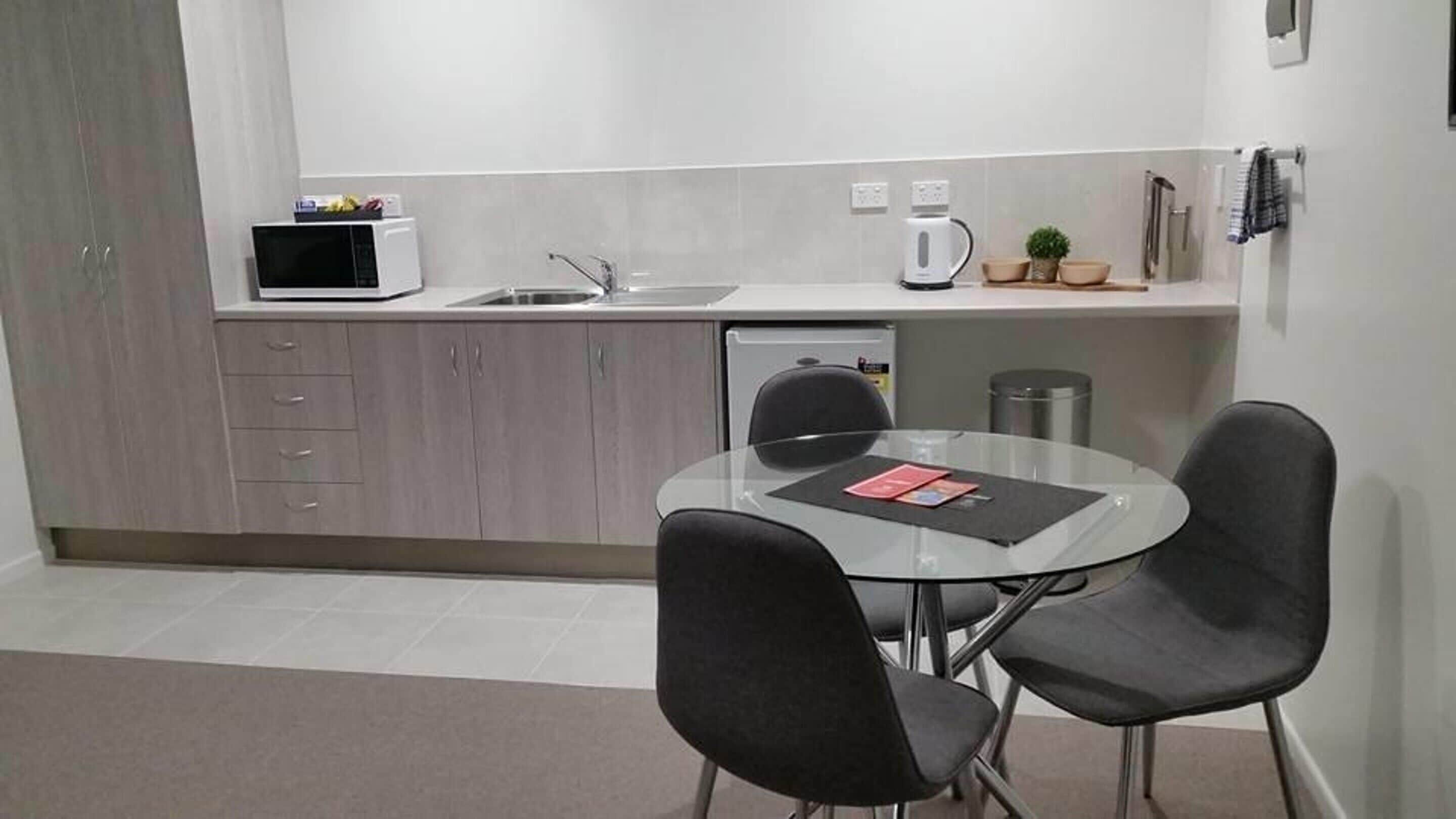 A compact kitchen with a microwave, coffee maker, and a small plant. There is a round glass table with two chairs and a few books.